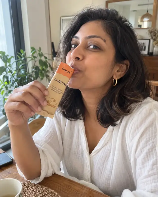 A woman drinking a LIVETAL sachet at a wooden table by the window